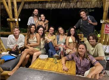 A group of eleven young adults, smiling and relaxed, sit together and pose at a wooden outdoor table under a thatched roof, enjoying a friendly gathering.