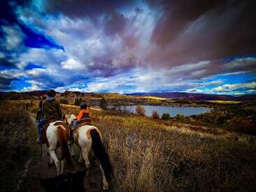 A group of people rides horses along a scenic trail, overlooking a serene lake and rolling hills under a dramatic, partly cloudy sky, surrounded by autumnal grass.