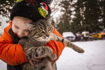 A child in an orange jacket and winter hat hugs a large tabby cat outdoors in the snow, trees and parked cars visible in the background.