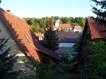 Das Bild zeigt eine ruhige Dorfszene mit einer Ansammlung von Häusern mit Terrakotta-Dächern, umgeben von üppigen Bäumen und einem Kirchturm in der Ferne.
