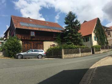 Das Bild zeigt eine ruhige Straßenszene mit einem zweistöckigen Haus mit rotem Dach, einem davor geparkten silbernen Auto und einem Holzzaun, der mit üppigem Grün geschmückt ist.