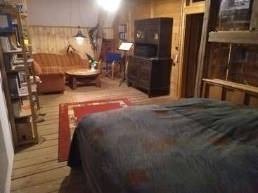 Das Bild zeigt ein gemütliches Zimmer mit einem Bett, einem Bücherregal und einer Couch, mit Holzböden und -wänden, einem roten Teppich und einem Fernsehgerät im Hintergrund.