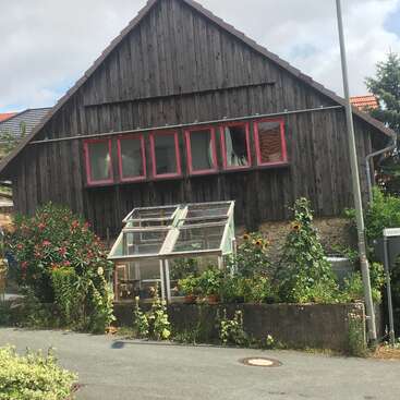 Das Bild zeigt ein rustikales Holzhaus mit einem schrägen Dach und einem Gewächshaus im Vorgarten, umgeben von üppigen Pflanzen und Blumen.