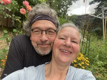 Das Bild zeigt ein Selfie von einem Mann und einer Frau, die vor einem Garten mit Blumen und Bäumen posieren. Beide lächeln und tragen legere Kleidung.