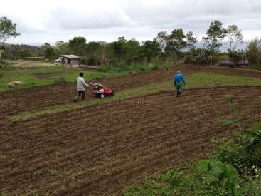 The image depicts two men tending to a field, one pushing a red rototiller and the other walking alongside, with a small shed and trees visible in the background.