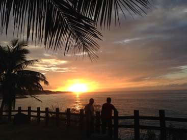 The image depicts two individuals standing by a fence, gazing out at a serene sunset over the ocean, with palm trees framing the tranquil scene.