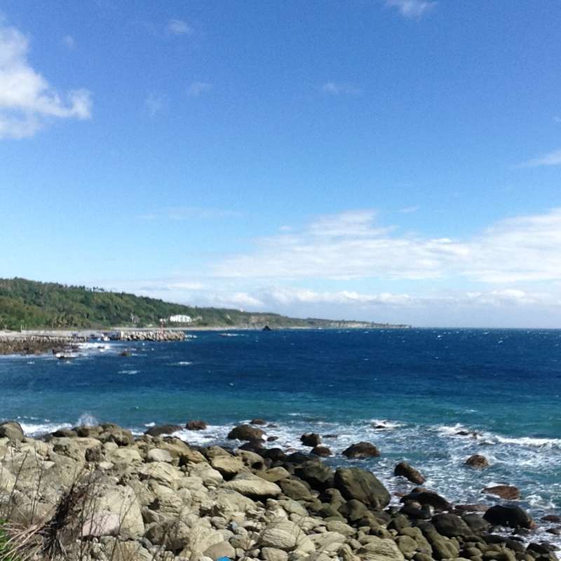 The image depicts a serene rocky coastline with a vibrant blue ocean, a distant pier, and a lush green hillside under a clear blue sky with white clouds.