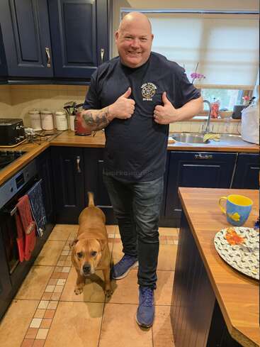 A smiling man in a navy shirt gives two thumbs up in a cozy kitchen, standing beside a brown dog. Blue cabinets and colorful dishes surround them.