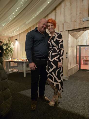 A smiling couple poses together in a warmly lit, rustic room with draped ceiling lights, wooden walls, floral arrangement, and a festive table in the background.