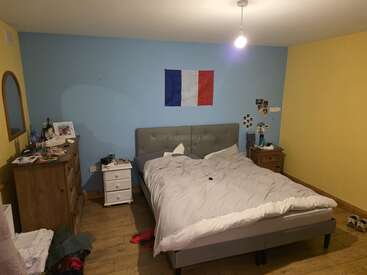 Une chambre confortable avec un lit double, des murs bleus et jaunes, un drapeau français au-dessus de la tête de lit, des meubles en bois, des vêtements éparpillés, un éclairage tamisé et des photos personnelles.