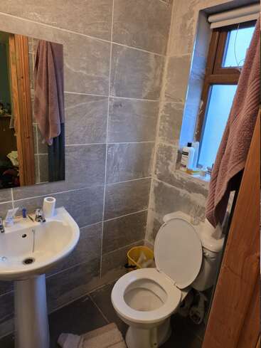 Cette image montre une petite salle de bains avec des murs carrelés gris, un lavabo sur pied, des toilettes avec le couvercle ouvert, une poubelle jaune, des serviettes et des articles de toilette.