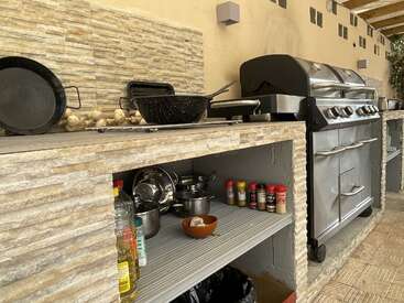 En esta imagen se ve una cocina exterior con una parrilla, utensilios de cocina, ollas, especias, aceite y ajo sobre una encimera de azulejos y estantes abiertos debajo.
