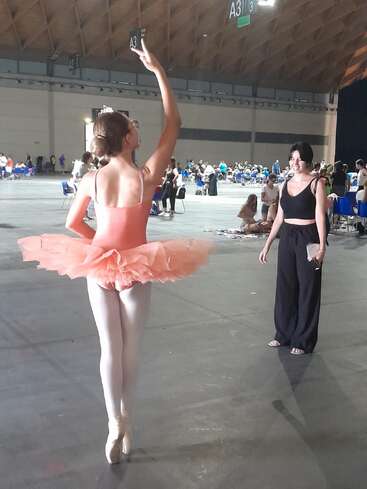 Une jeune ballerine vêtue d'un tutu rose prend une pose gracieuse sur pointes, tandis qu'une femme en noir l'observe, dans une grande salle spacieuse remplie de monde.