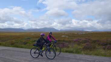 Deux personnes font du vélo sur une route de campagne, entourées de champs à perte de vue et de montagnes au loin, sous un ciel partiellement nuageux, profitant ensemble d'une belle balade en plein air.