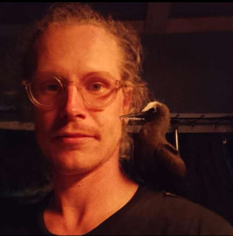 A man with glasses and curly hair is indoors, standing in dim lighting. A small dark bird with a white head rests comfortably on his shoulder.