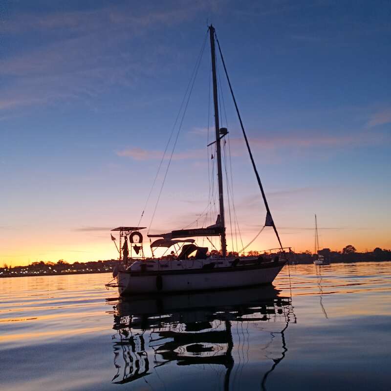 A sailboat floats peacefully on calm water at sunset, casting a beautiful reflection. The sky glows with pink, orange, and blue hues, creating tranquility.