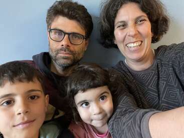 Una familia de cuatro miembros, entre ellos dos niños y dos adultos, está sentada muy junta. La mujer sonríe ampliamente, mientras que los demás tienen expresiones neutras. Ambiente informal.
