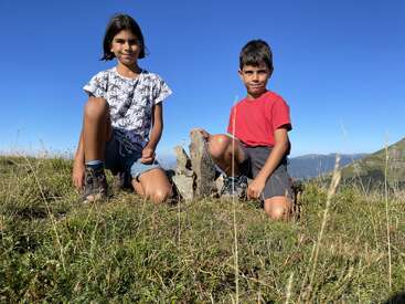 Dos niños se arrodillan en el suelo cubierto de hierba, sonriendo a la cámara. Están al aire libre, con montañas y cielo azul de fondo, y rocas dispuestas entre ellos.