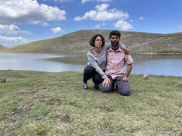 Un hombre y una mujer arrodillados juntos sobre la hierba cerca de un tranquilo lago. Las colinas y el cielo azul con nubes dispersas proporcionan un fondo natural tranquilo y pintoresco.