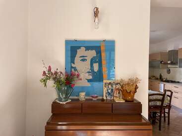 Un piano en bois contre un mur blanc présente des œuvres d'art, des fleurs dans des vases et divers objets décoratifs. Au-dessus, un portrait moderne et une ampoule apparente sont visibles. Cuisine à proximité.