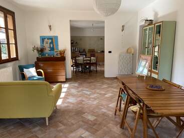 Salon-salle à manger lumineux et confortable avec des meubles en bois, de grandes fenêtres, des œuvres d'art et un canapé vert. La cuisine et l'armoire attenantes ajoutent du charme. Ambiance accueillante, aérée et familiale.