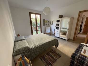 Une chambre confortable avec un lit double, une armoire, un fauteuil et des tapis colorés. Des portes-fenêtres s'ouvrent sur un jardin verdoyant. La décoration est simple, chaleureuse et accueillante.