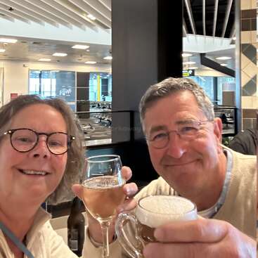 A smiling man and woman toast with drinks—wine and beer—in a bright, modern indoor setting, possibly a restaurant or airport lounge, enjoying their time together.
