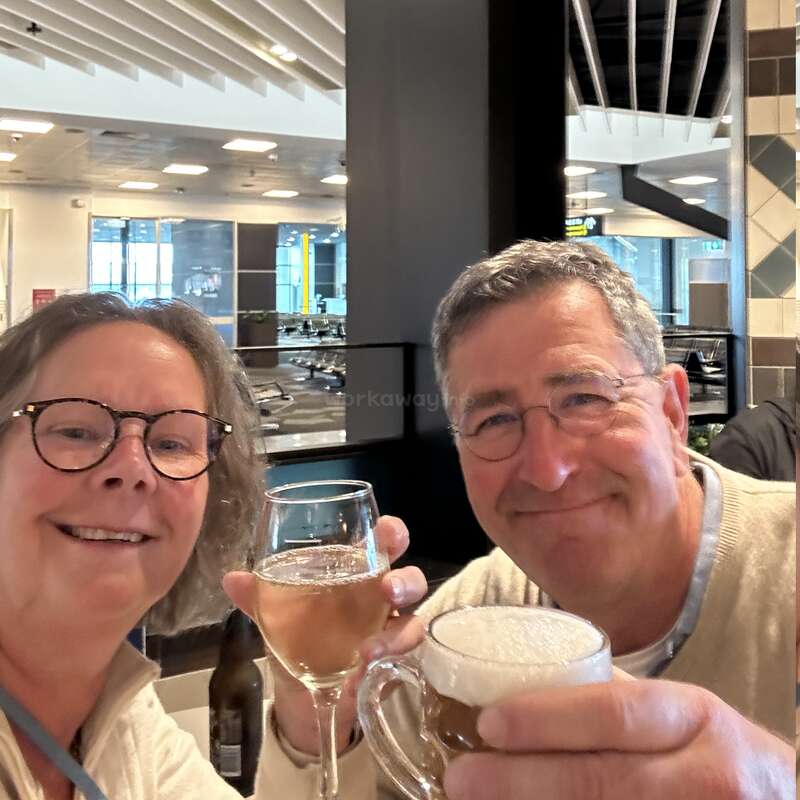A smiling man and woman toast with drinks—wine and beer—in a bright, modern indoor setting, possibly a restaurant or airport lounge, enjoying their time together.