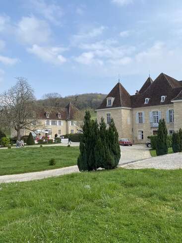 Das Bild zeigt ein großes Steinhaus mit steilen Dächern, umgeben von gepflegter Grünanlage, Wegen und Bäumen. Draußen spazieren Menschen unter einem teilweise bewölkten Himmel.