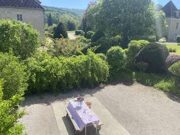 Auf einem bezaubernden Anwesen steht unter strahlend blauem Himmel ein wunderschön gedeckter Tisch mit einer lavendelfarbenen Tischdecke im Freien, umgeben von üppigen grünen Hecken und Bäumen.