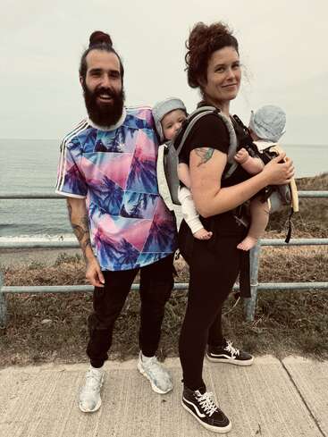 A cheerful couple stands together outdoors by the water, each carrying a baby in front carriers. The family looks happy and ready for a fun outing.