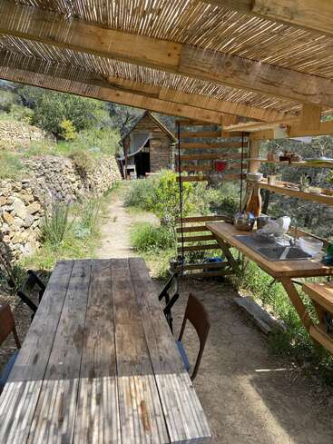 Eine rustikale Außenküche mit einem Holztisch und Stühlen, offenen Regalen und einer Spüle, umgeben von viel Grün und einem Steinweg, der zu einer kleinen Hütte führt.