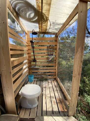 Dieses Bild zeigt ein Badezimmer im Freien mit Holzwänden, einer Toilette, einer Dusche, einem blauen Eimer und durchsichtigen Seitenwänden, umgeben von Bäumen und Sonnenlicht.