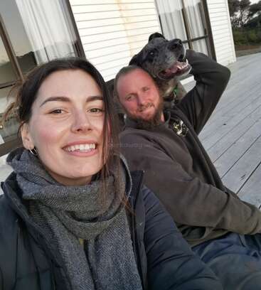 Eine Frau, ein Mann und ein glücklicher Hund sitzen auf einer Holzterrasse vor einem Haus, lächeln und genießen einen gemütlichen, entspannten Moment zusammen.