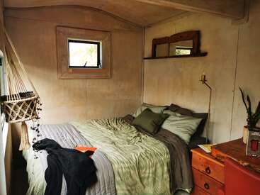 Dieses gemütliche Zimmer verfügt über ein ordentlich gemachtes Bett mit grüner Bettwäsche, ein kleines rustikales Fenster, einen hölzernen Wandspiegel, Topfpflanzen, einen Schreibtisch und hängende Pflanzendekoration.