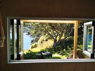 Ein großes Fenster gibt den Blick frei auf eine ruhige Landschaft mit üppigen Bäumen, Sonnenlicht, einer Holzterrasse, Grünpflanzen und einem friedlichen Fluss oder See im Hintergrund.