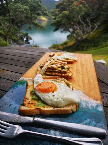 Auf einem Holzbrett liegt Toast mit Eiern, die im Freien serviert werden. Der malerische See, die Bäume und die Berge schaffen eine friedliche, einladende Frühstücksatmosphäre. Gabel und Messer liegen in der Nähe.