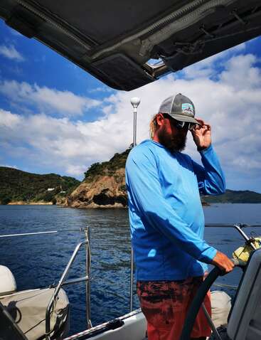 Ein Mann in blauem Hemd und Hut steht auf einem Boot und steuert, mit felsigen Inseln und blauem Himmel im malerischen Hintergrund, und genießt das sonnige Wetter.