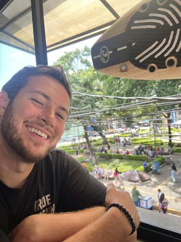 La imagen muestra a un hombre de pelo castaño corto y camiseta negra, sonriendo a la cámara, con una escena de parque visible a través de una ventana detrás de él.