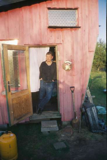 Das Bild zeigt eine Frau, die in der Tür eines kleinen, rosafarbenen Holzgebäudes mit einer offenen Tür und einem Fenster darüber steht, umgeben von Gartengeräten.