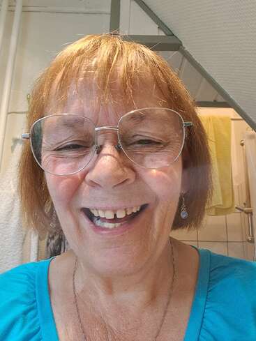 Une femme âgée souriante aux cheveux courts et auburn, portant de grosses lunettes et un haut turquoise, prend joyeusement un selfie dans une salle de bain lumineuse.