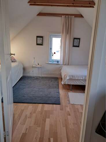Cette chambre mansardée confortable comprend deux lits simples, des planchers en bois, un tapis gris, une petite table d'appoint, une fenêtre avec rideau et des œuvres d'art murales minimalistes.