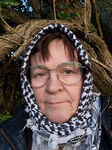 Une femme âgée portant de grosses lunettes et un foulard à motifs est assise à l'extérieur. Derrière elle, des branches d'arbre torsadées et des feuilles vertes créent un arrière-plan naturel et serein.