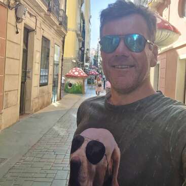 Ein Mann mit Sonnenbrille und einem T-Shirt mit einem Hund mit Sonnenbrille steht lächelnd auf einer sonnigen Straße, die von Gebäuden und skurrilen Pilzdekorationen im Hintergrund gesäumt ist.