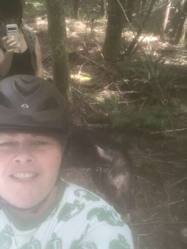Zwei Personen befinden sich in einem Wald. Die Person vorne trägt einen Helm und lächelt für ein Selfie, während eine andere ein Foto macht. Ein Pferd steht in der Nähe.