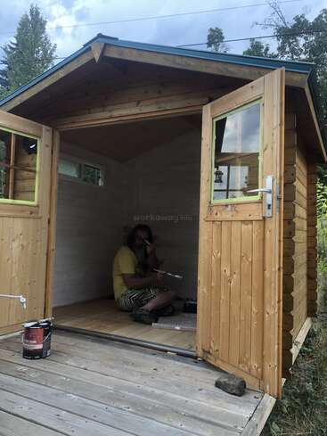 A person sits inside a small wooden shed with open doors, painting the interior white. Paint cans and brushes are visible on the wooden porch outside.