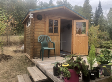 A small wooden garden shed with a green plastic chair on its porch. Potted plants decorate the steps. Trees and shrubs surround the shed. Serene setting.
