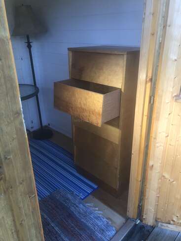 Auf dem Bild sieht man ein kleines gemütliches Zimmer mit einer Kommode aus Holz, einer offenen Schublade, einer Stehlampe, einem runden Beistelltisch und zwei gewebten Teppichen.