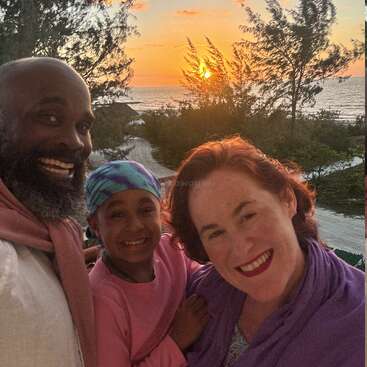 A smiling family of three enjoys a beautiful beach sunset, with trees, sandy paths, and the ocean in the background, capturing a joyful vacation moment together.