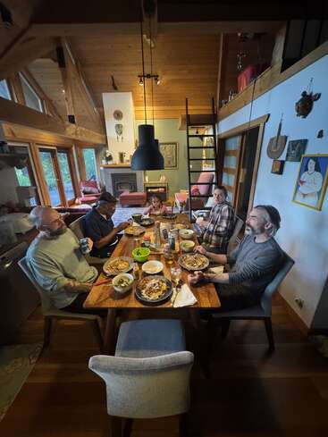 Five people are gathered around a wooden dining table in a cozy, modern cabin, sharing a meal together. Warm lighting highlights the inviting atmosphere.